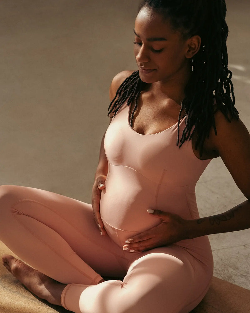 Pregnant woman holding her belly in a pink outfit sitting on a beige surface.